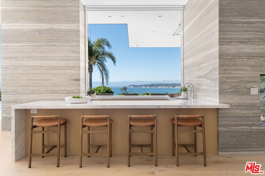 This sophisticated kitchen features a sleek, marble-topped island with four wooden bar stools, set against a backdrop of striking, horizontally-veined stone walls. A large, floor-to-ceiling window frames a breathtaking, unobstructed view of the ocean and coastline, seamlessly blending indoor luxury with the natural landscape. The minimalist design and warm wood tones create an airy, high-end atmosphere perfect for coastal living.