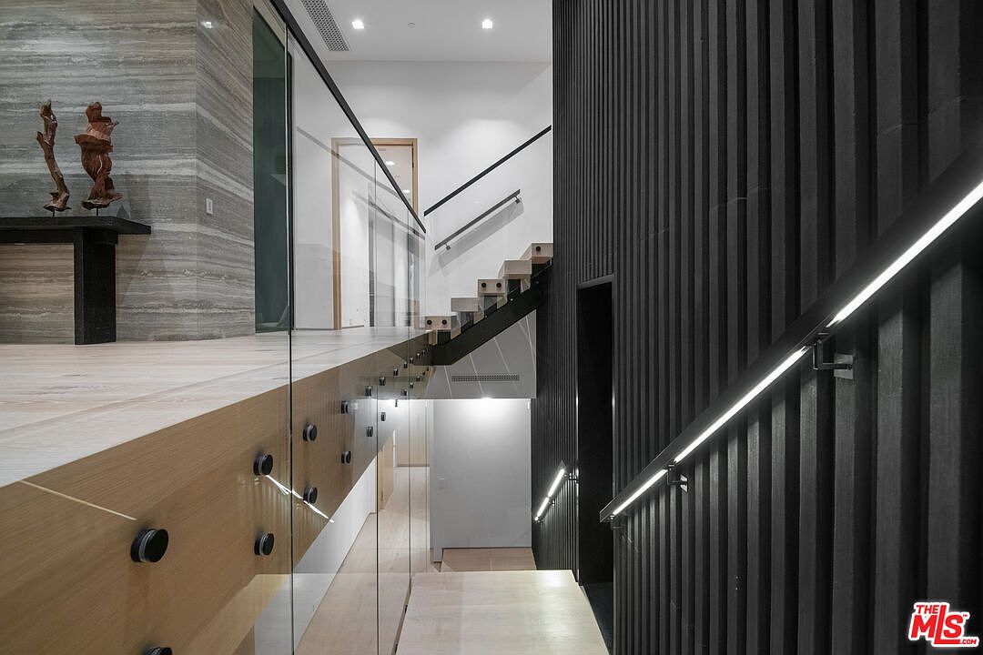 This sophisticated interior space features a dramatic staircase design with a glass railing and a striking black slatted accent wall illuminated by integrated LED handrail lighting. The perspective looks down the staircase, highlighting the contrast between the light wood flooring and the dark, modern architectural elements. The clean lines and high-end materials create a luxurious, contemporary atmosphere typical of a high-end modern residence.