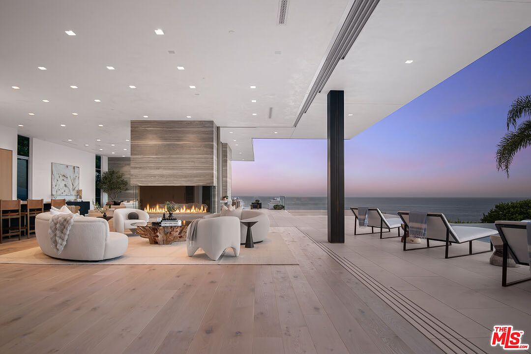 This expansive, open-concept living room seamlessly blends indoor and outdoor living through massive floor-to-ceiling sliding glass doors that open to a stunning oceanfront deck. The space features a striking, central stone-clad fireplace, light hardwood flooring, and contemporary, plush white furniture that creates a sophisticated and airy atmosphere. The perspective captures the transition from the luxurious interior lounge area to the serene twilight horizon over the sea, emphasizing the home's premium coastal location.