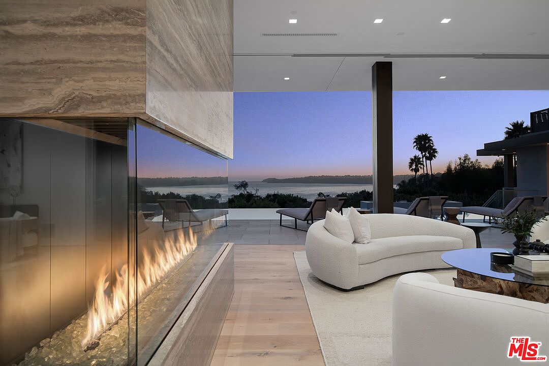 This sophisticated living room features a sleek, modern fireplace with a glass enclosure that serves as a focal point, seamlessly blending indoor and outdoor living. The space is furnished with a plush, curved white sofa and opens directly onto a spacious patio with stunning sunset views over the ocean. The minimalist design, high ceilings, and neutral color palette create an atmosphere of luxury and tranquility.