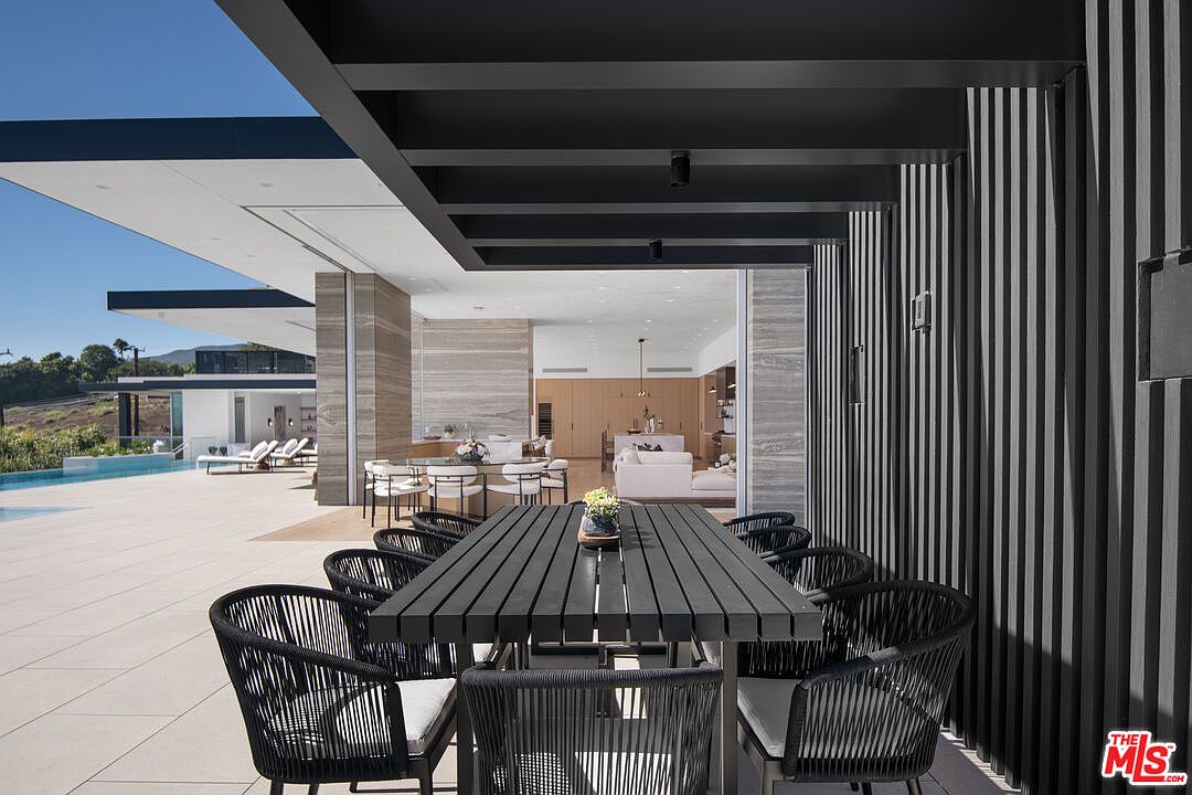 This sophisticated outdoor dining area features a sleek, black slatted dining table surrounded by matching woven chairs, set beneath a modern covered patio with dramatic vertical black wall paneling. The space seamlessly transitions into an expansive tiled pool deck, offering a clear view of the open-concept interior living area and a stunning infinity-edge pool. The architectural design emphasizes a luxurious, contemporary aesthetic with clean lines and a harmonious indoor-outdoor flow.