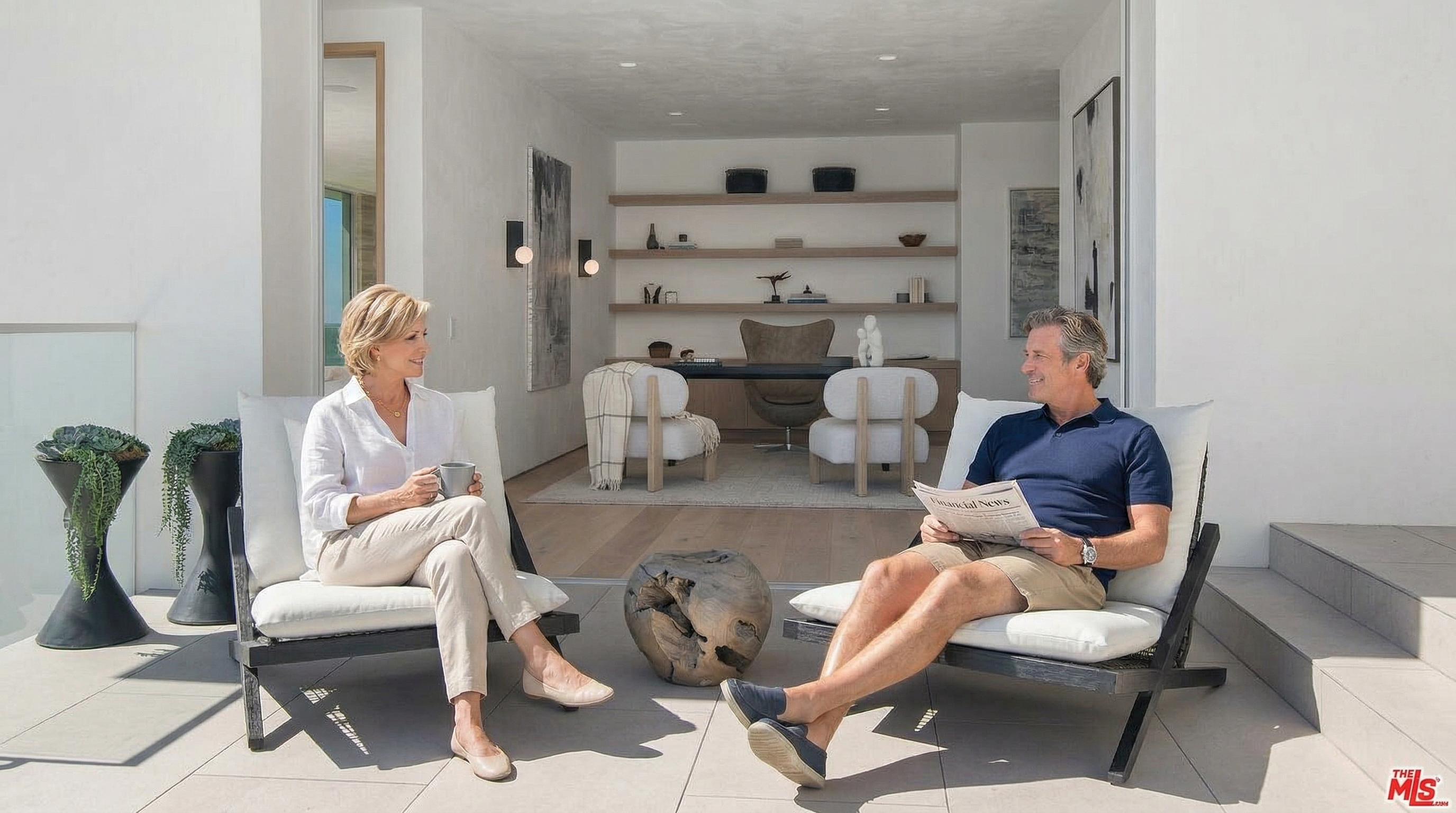 This modern outdoor patio space features sleek, light-toned stone tiling and two contemporary lounge chairs positioned near a sparkling pool edge. The patio seamlessly transitions into an indoor office or study area through a wide, open doorway, showcasing a minimalist design with built-in shelving and a desk. The overall aesthetic is luxurious and airy, emphasizing a sophisticated indoor-outdoor lifestyle.