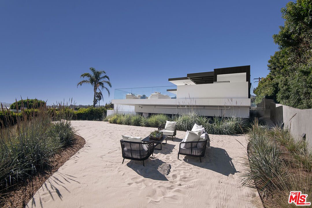 This outdoor space features a unique, sandy ground surface designed to mimic a beach, complete with a modern patio furniture set arranged for relaxation. The area is framed by lush, ornamental grasses and overlooks a contemporary, multi-level architectural home with a glass-railed balcony. The bright, clear blue sky and the presence of a tall palm tree contribute to a serene, coastal-luxury atmosphere.