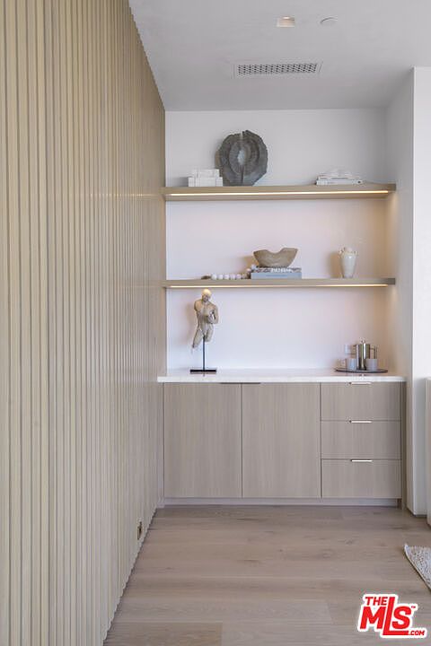 This image showcases a sophisticated built-in wet bar or display area featuring light wood cabinetry and matching floating shelves with integrated LED lighting. The wall is accented with vertical wood slats, creating a modern, textured aesthetic that complements the minimalist decor and neutral color palette. The perspective is a straight-on, eye-level shot that highlights the clean lines and high-end finishes of this functional living space.