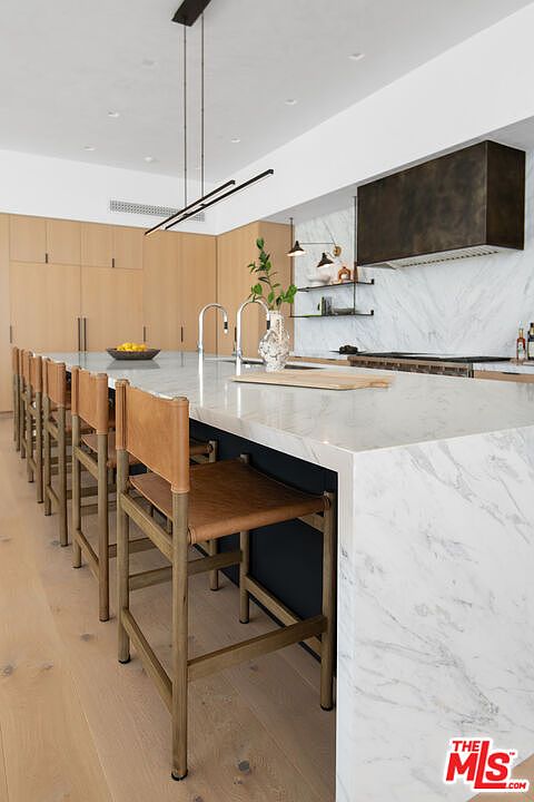 This sophisticated, modern kitchen features a large waterfall-edge marble island paired with rustic leather-seated bar stools. The space is defined by sleek light wood cabinetry, a dramatic dark metal range hood, and minimalist pendant lighting, creating a high-end, contemporary aesthetic. The perspective captures the length of the island, emphasizing the clean lines and premium materials that define this luxury culinary space.