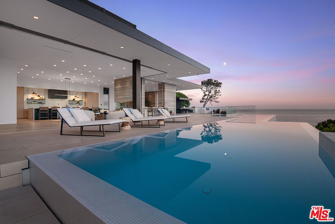 This stunning contemporary luxury home features a seamless indoor-outdoor transition, showcasing a sleek infinity-edge pool that overlooks the ocean at sunset. The expansive patio is furnished with modern lounge chairs, creating an inviting space for relaxation under a soft, twilight sky. The architectural design emphasizes clean lines, open-concept living, and a sophisticated, high-end aesthetic.