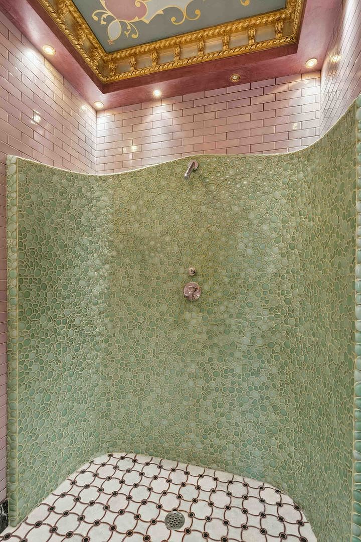 This unique shower features a curved wall finished in textured, light green pebble-style tile that creates a distinct organic aesthetic. The shower floor is lined with a decorative white tile featuring a repeating circular pattern, while the surrounding walls are clad in soft pink subway tile. Above, a recessed ceiling panel is framed by ornate gold molding, adding a touch of classical elegance to the space.