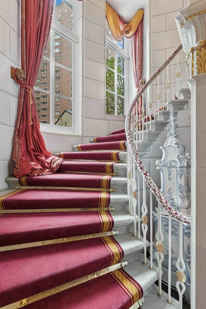 This elegant staircase features a grand, curved design with white stone steps adorned by a rich red runner with gold trim. The ornate white metal railing is accented with gold detailing, and the walls are finished with classic stone paneling. Tall windows draped in luxurious red and gold curtains allow natural light to illuminate the space, creating a sophisticated and opulent atmosphere.