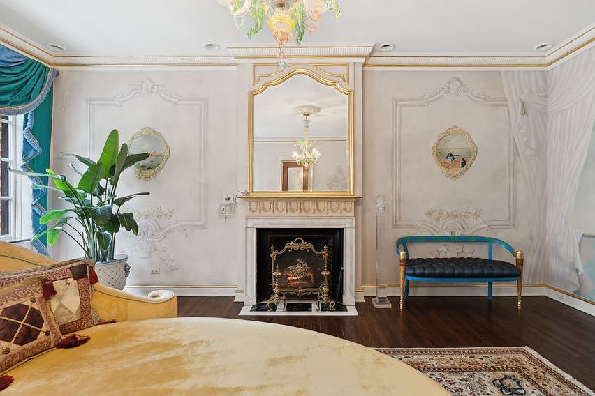 This opulent bedroom features a central fireplace with an ornate, tall mantel and a large, gold-framed mirror above it. The walls are adorned with decorative molding and hand-painted murals, complemented by a plush, velvet-upholstered bench and a large potted plant. The room is anchored by a dark wood floor and a patterned area rug, creating a sophisticated and luxurious atmosphere.