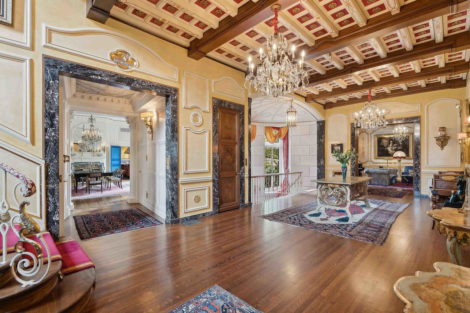 This grand foyer showcases an opulent, traditional design with a coffered ceiling featuring dark wood beams and decorative panels. The space is anchored by a polished wood floor, a central console table, and multiple crystal chandeliers that cast a warm glow across the room. Through the arched openings, one can see into adjacent formal living and dining areas, emphasizing the home's expansive and interconnected layout.