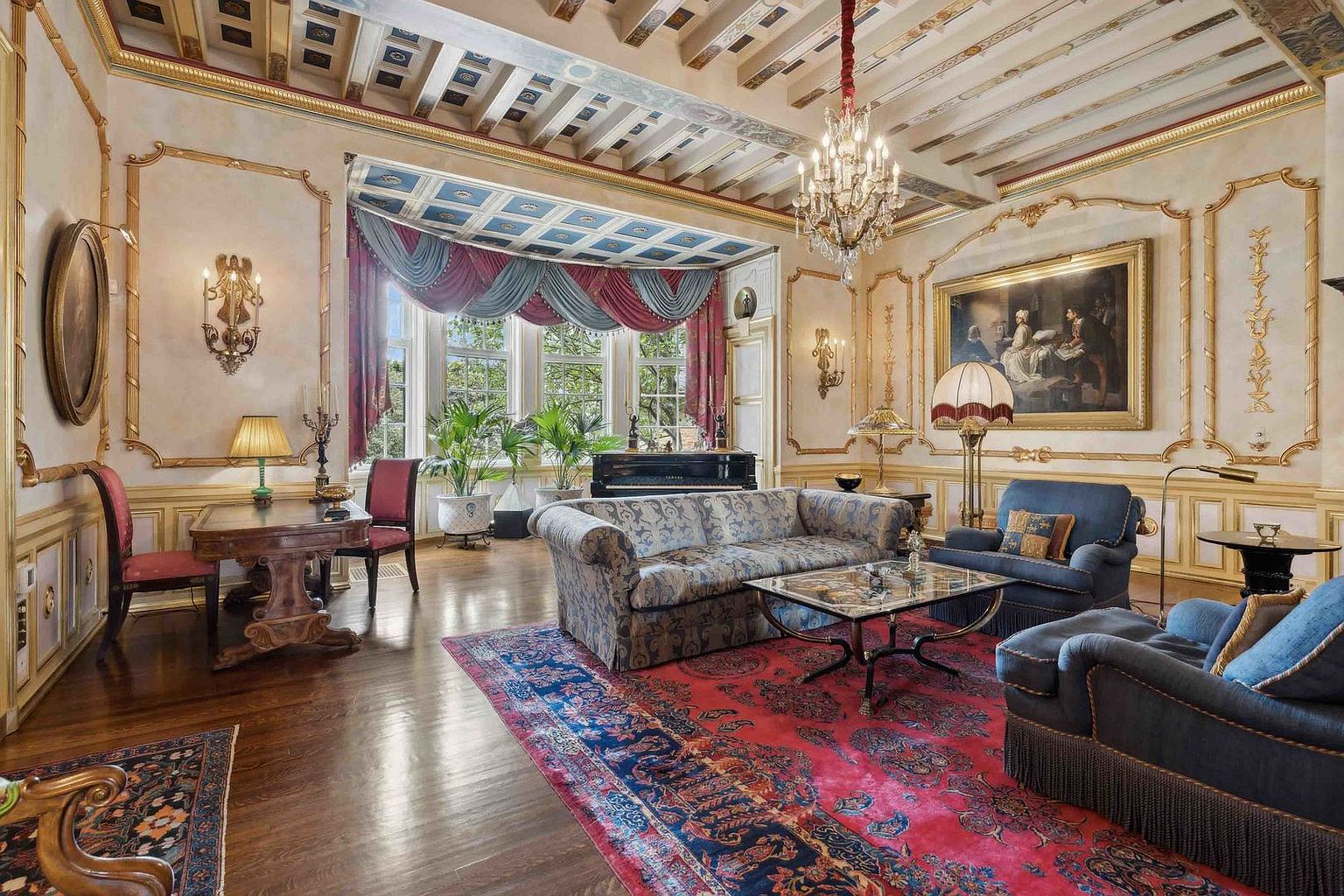 This opulent living room features ornate gold-trimmed wall moldings, a coffered ceiling with decorative painting, and a grand crystal chandelier. The space is furnished with a patterned sofa, plush blue armchairs, and a central glass-topped coffee table, all resting on a large, vibrant red area rug. A bay window area with elegant drapery frames a view of the outdoors, while a piano and a writing desk add to the sophisticated, classic aesthetic.