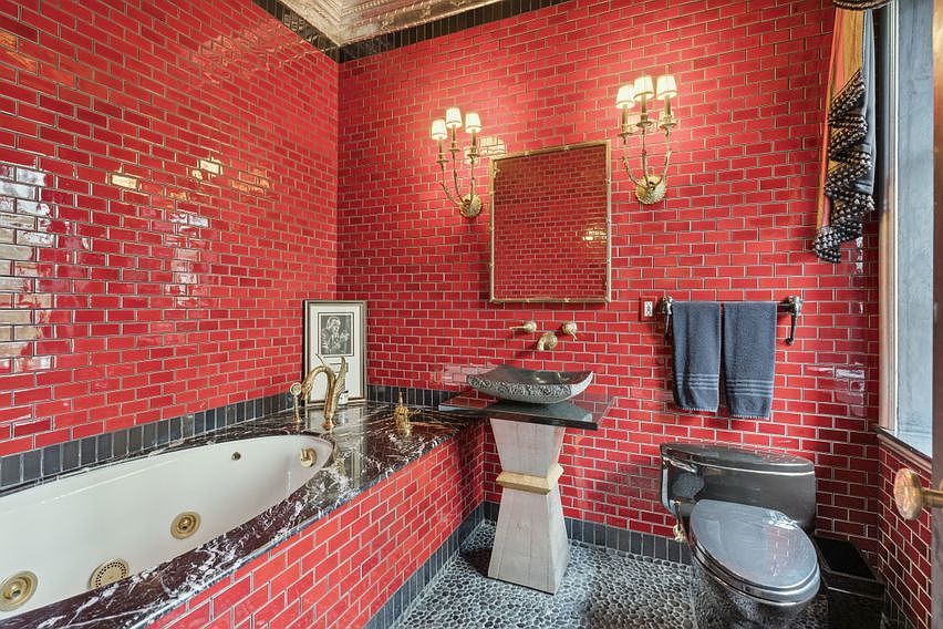 This bold bathroom features striking red subway tile walls contrasted by dark stone accents and a pebble-textured floor. The space includes a jetted soaking tub with a dark stone surround, a unique stone vessel sink perched on a pedestal, and a modern toilet. Two ornate wall sconces flank a rectangular mirror, creating a dramatic and high-contrast aesthetic.
