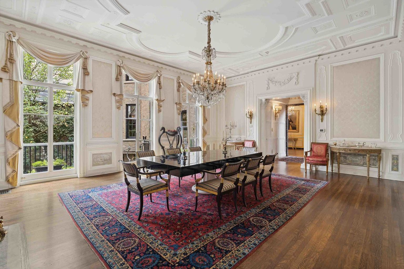 This elegant formal dining room features a large, dark-finished table surrounded by upholstered chairs, all set atop a vibrant, patterned area rug. The space is defined by ornate crown molding, tall windows draped in soft fabric, and a grand crystal chandelier that serves as a central focal point. The room exudes a sophisticated, traditional aesthetic with light-toned walls and polished wood flooring, offering a view into an adjacent hallway.