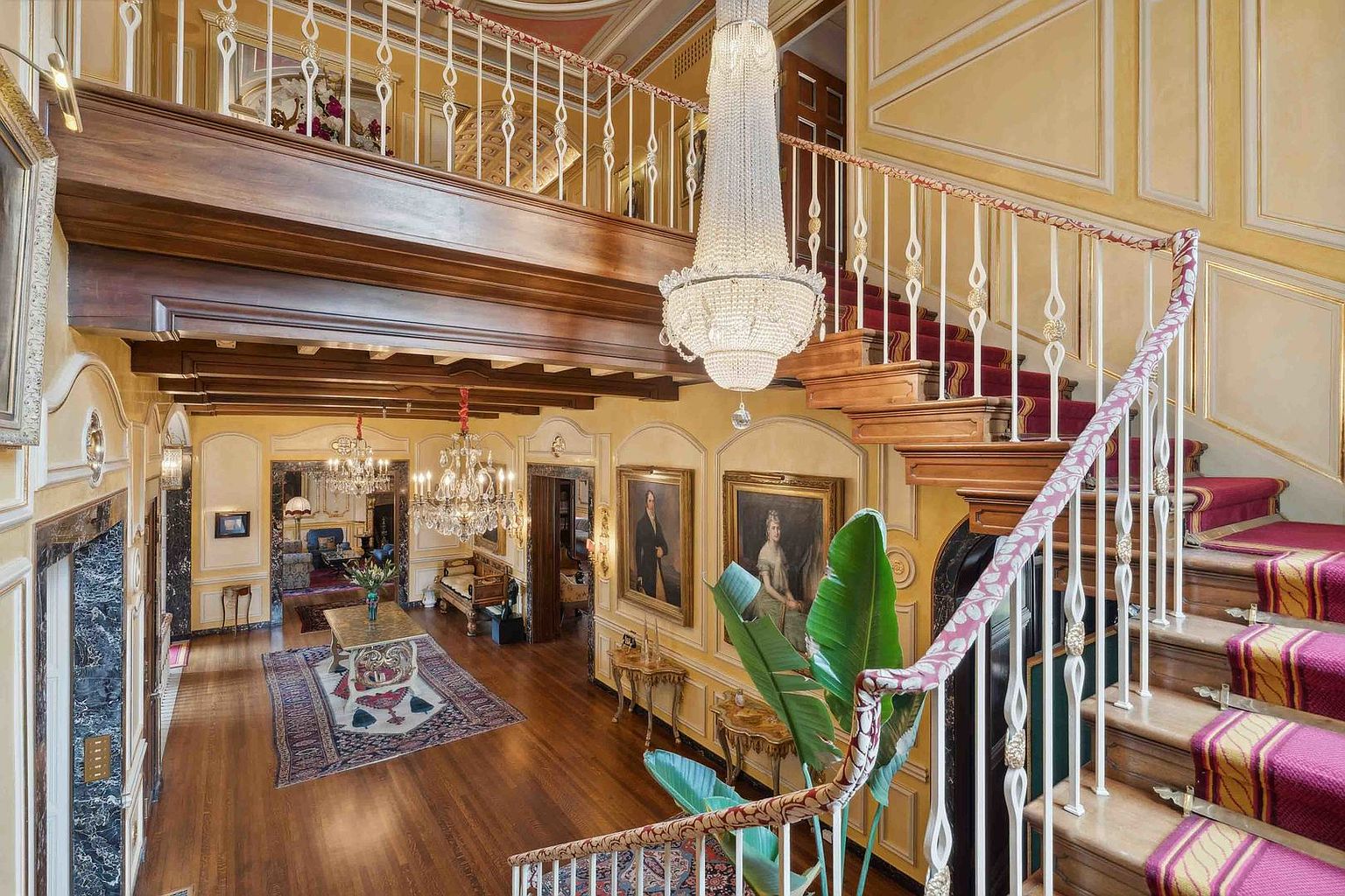 This grand foyer features a sweeping staircase with a patterned runner and ornate metal railings, leading to an upper-level gallery. A massive crystal chandelier serves as the focal point, illuminating the polished wood floors and the formal living area below. The space is decorated with classic portrait paintings and elegant furnishings, creating a sophisticated and opulent atmosphere.