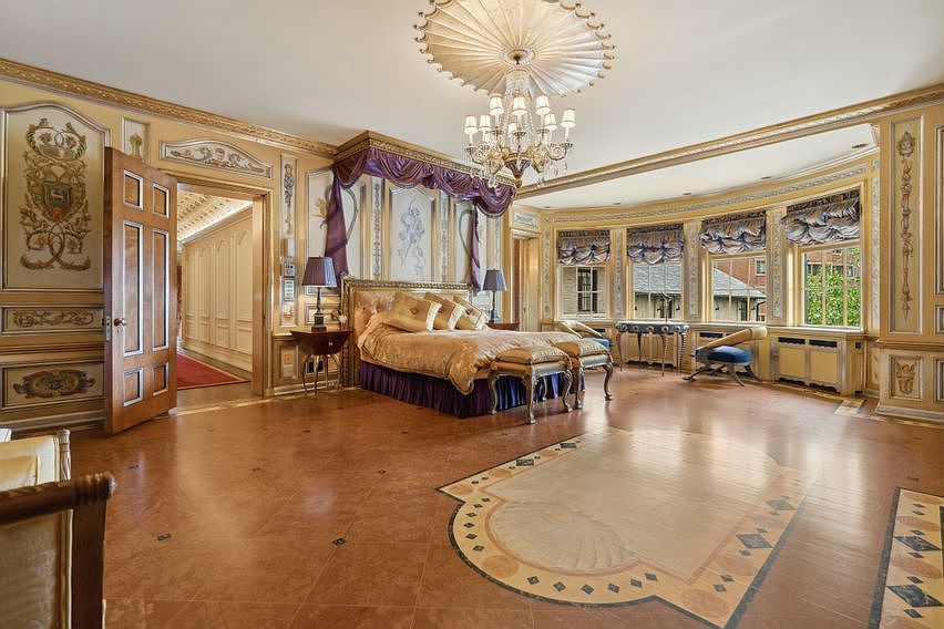This opulent primary bedroom features an ornate, classical design with intricate wall moldings, a grand crystal chandelier, and a canopy bed as the central focal point. The room is finished with patterned tile flooring that includes a decorative inlay, and a curved wall of windows draped in elegant fabric valances provides natural light. The perspective is wide-angle, capturing the luxurious scale of the space and the transition into an adjacent hallway.