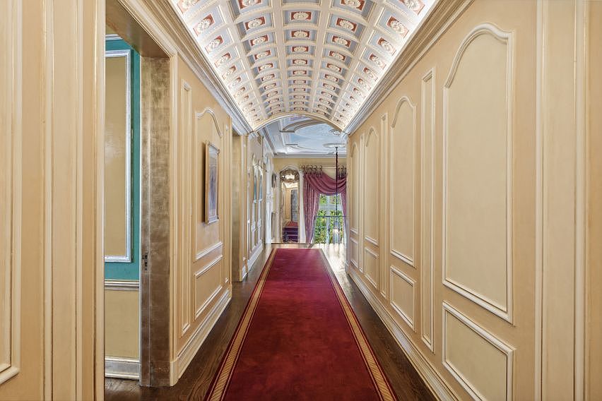 This elegant, formal hallway features a dramatic barrel-vaulted ceiling adorned with intricate, light-toned coffered paneling. A long, deep red runner stretches across the dark wood flooring, leading the eye toward a bright, arched window at the far end draped in soft purple curtains. The walls are finished with classic wainscoting and molding, creating a sophisticated, gallery-like atmosphere.