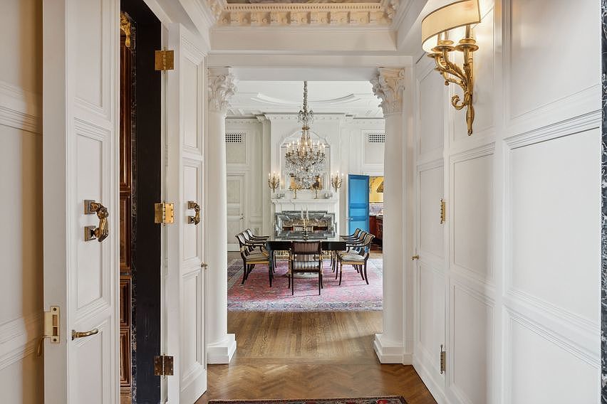 This perspective captures a grand transition space, looking from a paneled hallway through a pair of open doors into a formal dining room. The foreground features classic white wainscoting and ornate brass hardware, while the dining area beyond showcases a crystal chandelier, a dark wood table, and a fireplace with a decorative stone surround. The space is defined by its classical architectural details, including columns and intricate crown molding, creating an atmosphere of refined elegance.