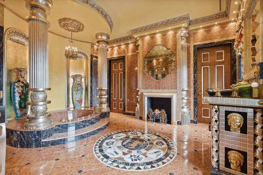This opulent primary bathroom features a sunken soaking tub set within a raised platform, framed by decorative columns and large, ornate vases. The room is finished with polished stone flooring, a central circular mosaic inlay, and a fireplace with a tall, decorative surround and mirror above. The space is characterized by its dramatic, classical-inspired design, warm lighting, and rich, textured surfaces.