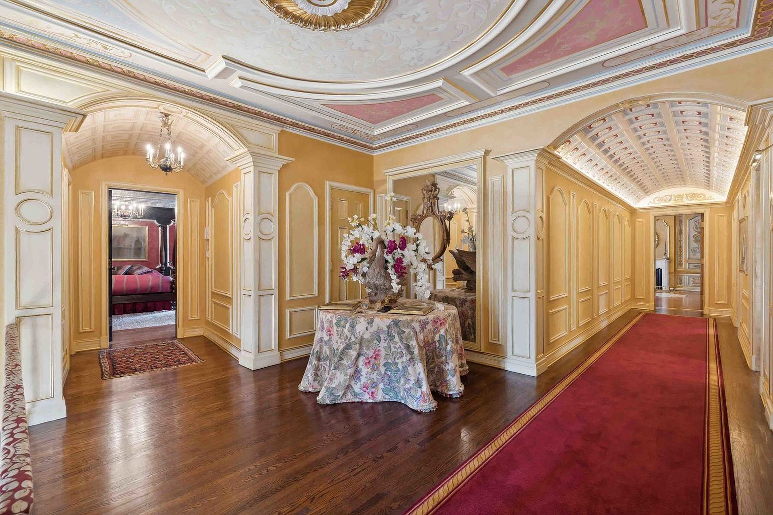 This elegant, formal hallway features a grand aesthetic with warm yellow walls, intricate crown molding, and a barrel-vaulted ceiling. A long red runner stretches down the corridor, leading the eye toward a distant doorway, while a central round table with a floral tablecloth serves as a focal point beneath a decorative mirror. The perspective is centered, capturing the symmetrical architectural details and the transition into an adjacent bedroom space.