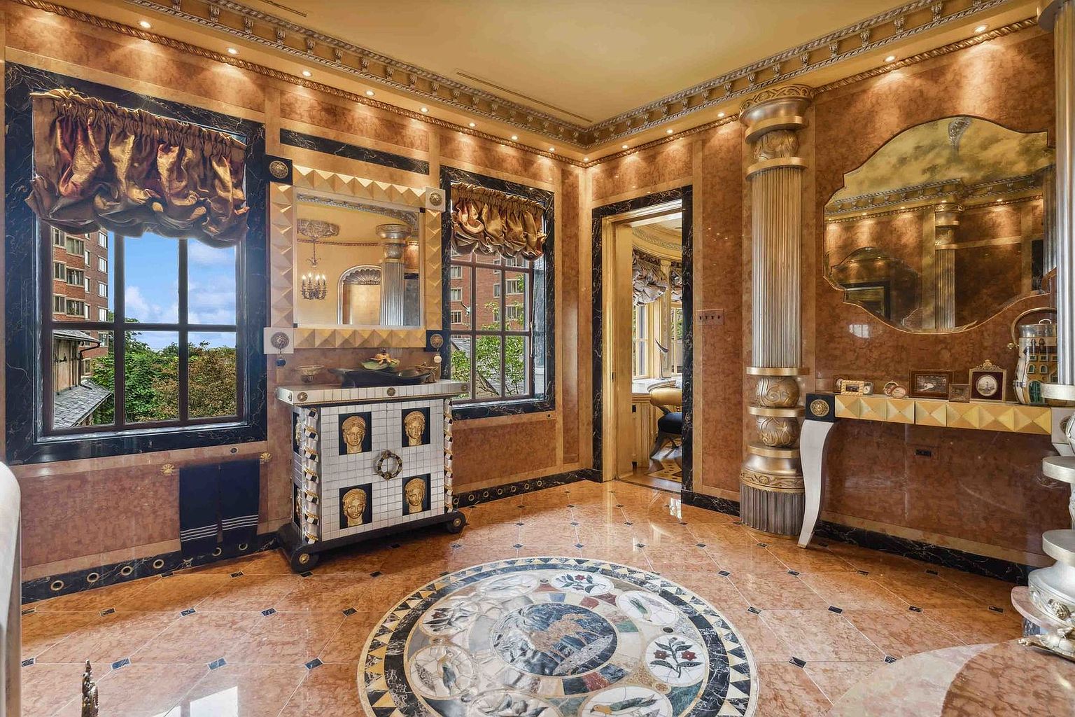 This opulent bathroom features a dramatic, classical aesthetic with rich, warm-toned stone wall paneling and a central circular mosaic floor inlay. A vanity cabinet adorned with gold-toned face motifs sits between two windows, while a prominent metallic column adds a grand architectural element to the space. The room is finished with ornate crown molding and decorative mirrors, creating a luxurious and highly stylized atmosphere.