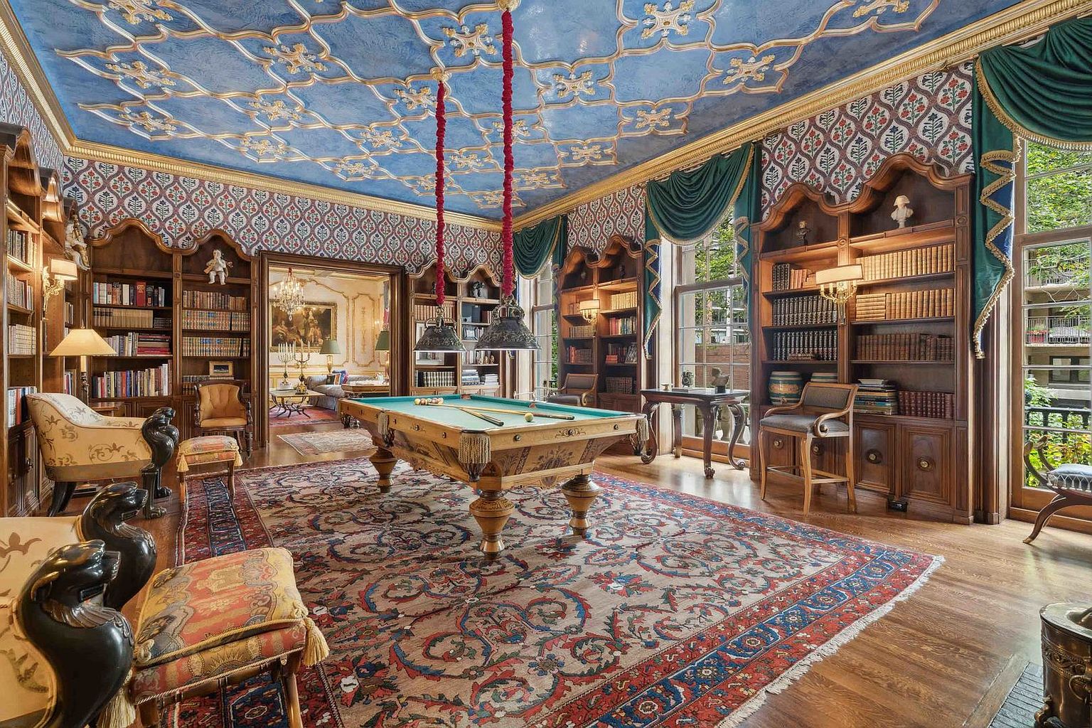 This opulent library and game room features a central billiard table set beneath ornate hanging lights, surrounded by extensive custom wood shelving filled with books. The room is defined by its dramatic blue ceiling with gold-toned decorative molding, patterned wallpaper, and a large, intricate area rug covering the hardwood floors. Large windows draped in heavy teal curtains provide natural light, while the overall aesthetic exudes a classic, sophisticated, and historic atmosphere.