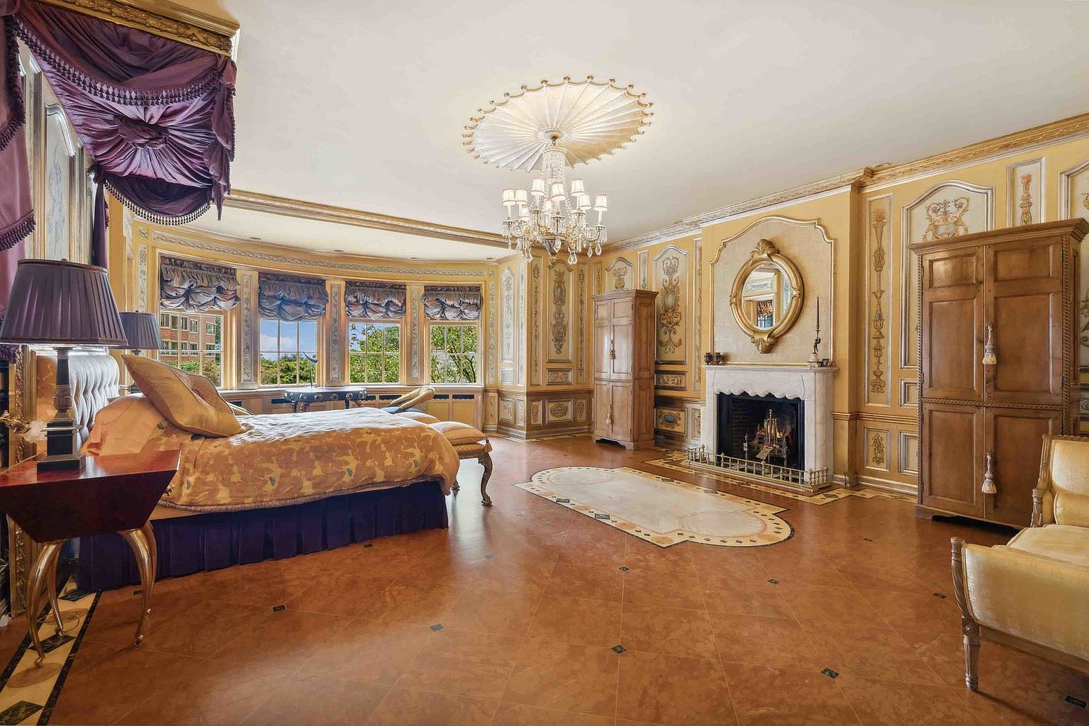This opulent primary bedroom features ornate gold-toned wall paneling, a crystal chandelier, and a large bed draped in rich purple fabric. A marble fireplace with a decorative mirror above serves as a focal point, while a curved wall of windows provides natural light and views of the outdoors. The room is finished with patterned tile flooring and a central rug, creating a sophisticated and regal atmosphere.