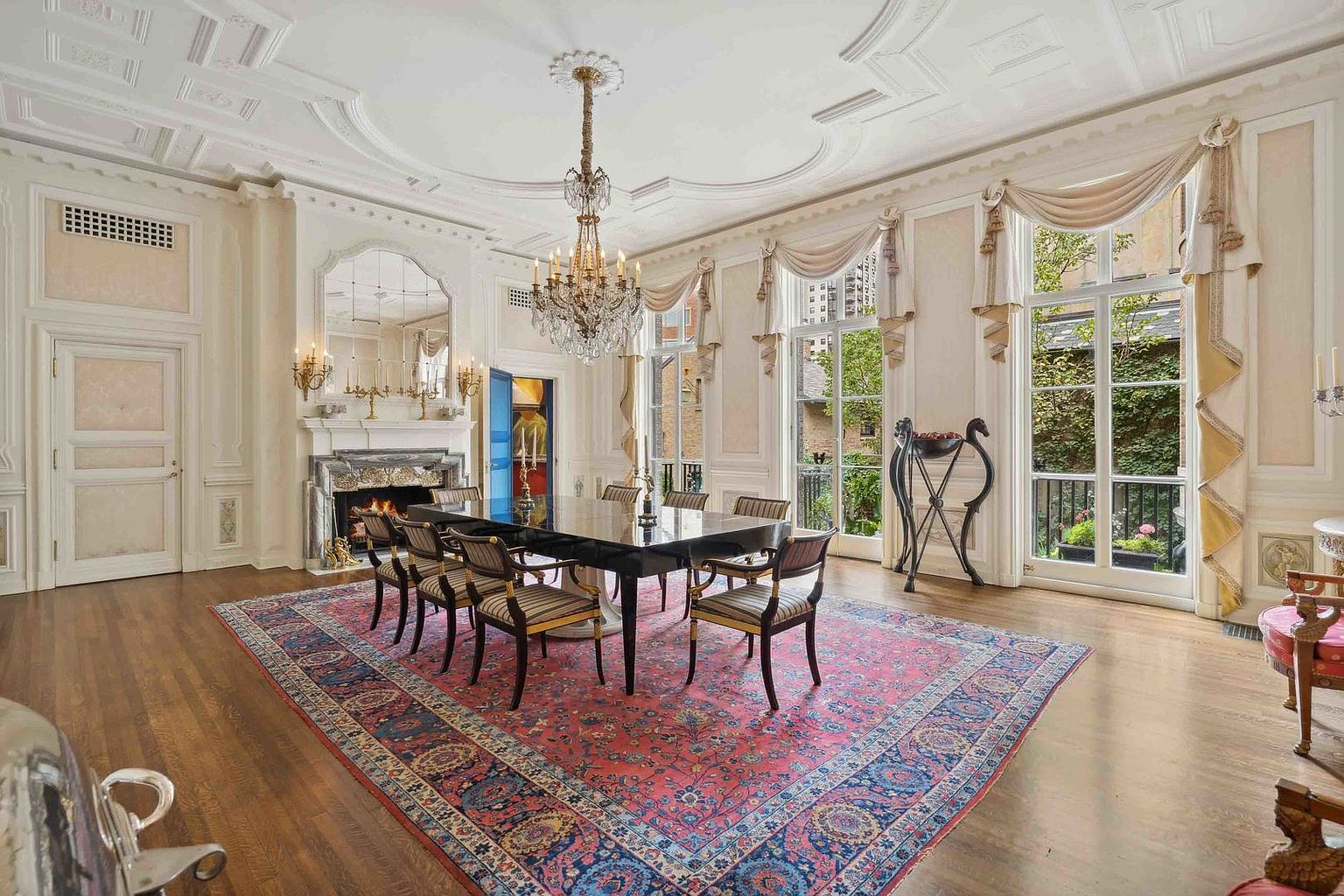 This elegant formal dining room features a large, dark-finished dining table surrounded by striped upholstered chairs, set atop a vibrant, intricately patterned area rug. The space is defined by ornate crown molding, a crystal chandelier, and a stone fireplace with a decorative mirror above it. Tall windows with draped valances allow natural light to fill the room, creating a sophisticated and classic atmosphere.