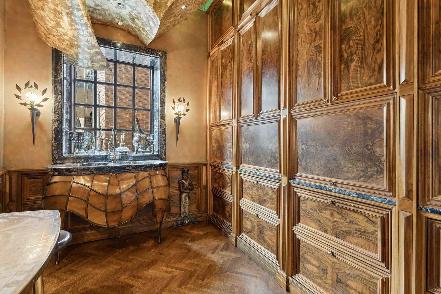 This opulent room features intricate wood paneling with a rich, swirling grain pattern covering the walls and built-in cabinetry. A unique, curved vanity with a dark stone top and metal lattice detailing sits beneath a window, flanked by ornate wall sconces. The space is finished with a herringbone-patterned wood floor and a dramatic, textured ceiling treatment, creating a sophisticated and highly detailed atmosphere.