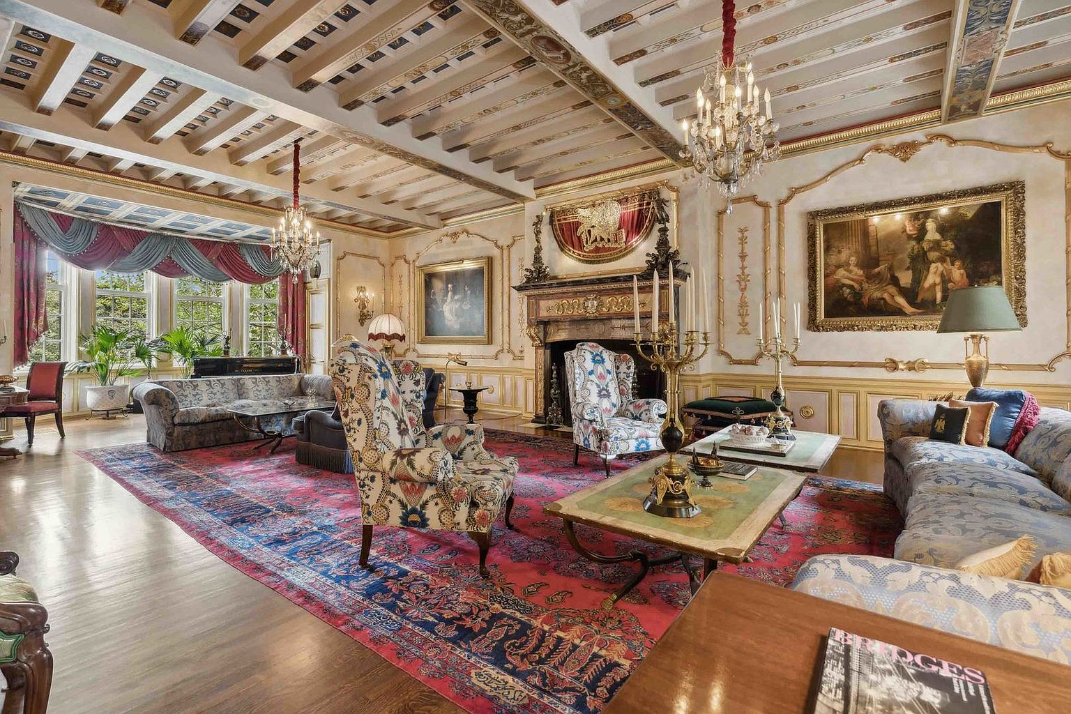 This opulent living room features a grand, traditional aesthetic with intricate crown molding, a coffered ceiling, and a large, ornate fireplace with a decorative mantel. The space is furnished with plush, patterned armchairs and sofas arranged on a vibrant, large-scale area rug, all illuminated by elegant crystal chandeliers. The perspective captures the expansive room from a slightly elevated angle, highlighting the sophisticated decor and the natural light streaming through the large windows.