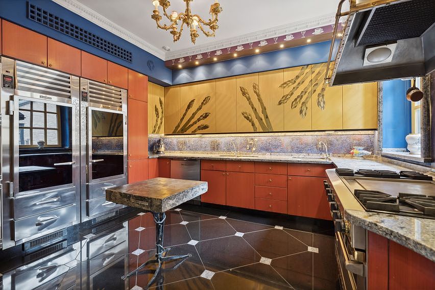 This unique kitchen features a bold design with vibrant orange cabinetry, a striking asparagus-themed mural on the upper cabinets, and a dark, geometric-patterned tile floor. A central butcher-block island with a sculptural metal base stands in the foreground, while professional-grade stainless steel appliances, including a large refrigerator and a range, define the workspace. The room is illuminated by an ornate gold chandelier and recessed lighting, creating a dramatic and highly personalized culinary space.