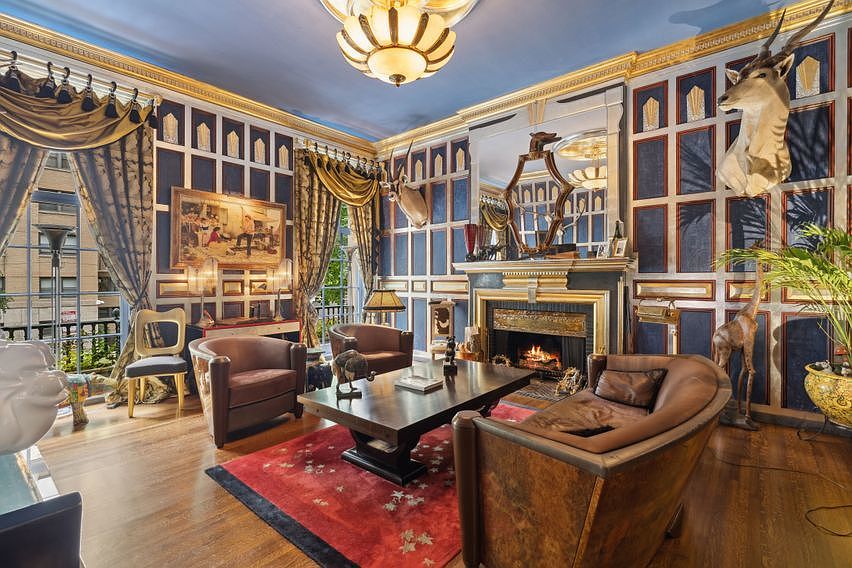 This opulent living room features deep blue paneled walls with gold trim, creating a sophisticated and dramatic atmosphere. The space is furnished with leather armchairs and a curved sofa arranged around a central coffee table, all set upon a vibrant red patterned rug. A prominent fireplace with a decorative mantel serves as a focal point, complemented by taxidermy accents and a large, ornate chandelier hanging from the ceiling.
