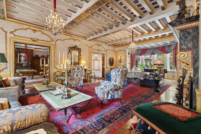 This opulent living room showcases a grand, traditional aesthetic with intricate gold-leaf detailing on the walls and a coffered wood-beam ceiling. The space is anchored by a large, vibrant red patterned rug, multiple ornate armchairs, and a central coffee table, while a glimpse into an adjacent room reveals a billiards table. The room is illuminated by elegant crystal chandeliers and features tall windows draped in layered fabric, creating a sophisticated and historic atmosphere.