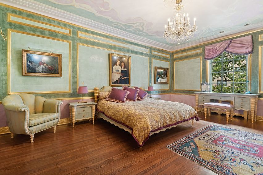 This elegant primary bedroom features ornate wall paneling with gold trim, a hand-painted ceiling mural, and a crystal chandelier. The room is furnished with a bed dressed in a patterned gold coverlet, a plush armchair, and a vanity desk positioned beneath a large window with draped valances. A traditional patterned area rug sits on the dark wood flooring, creating a sophisticated and classic atmosphere.