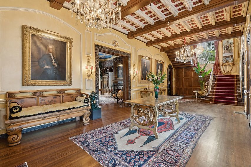 This grand foyer showcases an opulent, traditional design with a coffered wood-beam ceiling and a large crystal chandelier. A long, ornate wooden bench sits beneath a large framed portrait, while a central table rests on a detailed patterned rug. The space leads toward a staircase with a red runner, creating a sophisticated and welcoming entrance.