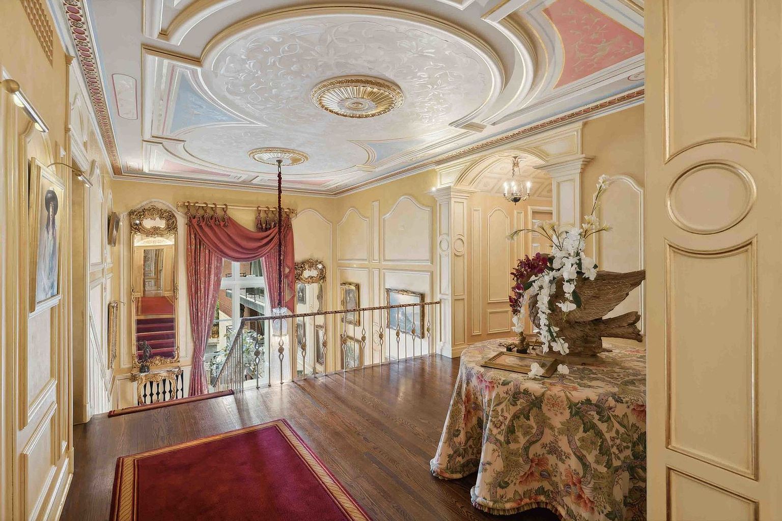 This elegant upper-level landing showcases ornate neoclassical design with intricate ceiling molding, soft pastel accents, and gold-leaf detailing. A round table draped in a floral tablecloth sits in the foreground, while a wrought-iron railing overlooks the staircase below. The space is framed by warm, neutral wall paneling, a large decorative mirror, and a rich red runner, creating a sophisticated and opulent atmosphere.