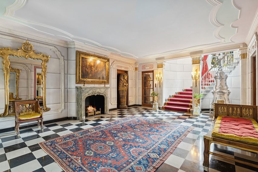 This opulent grand foyer or living space features a striking black-and-white checkered floor and an intricate, ornate ceiling design. The room is anchored by a stone fireplace with a large, gilded-frame painting above it, flanked by a decorative Egyptian-style sarcophagus and a massive, elaborate gold-framed mirror. A grand staircase with a red runner leads to an upper level, while a plush bench and a large, patterned area rug complete the luxurious, classical aesthetic.