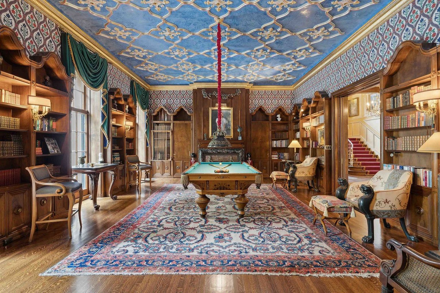 This opulent game room features a central billiard table set upon a large, intricate area rug, surrounded by extensive wood-paneled walls lined with built-in bookshelves. The room is distinguished by a striking blue ceiling with ornate gold-toned molding and a hanging pendant light, complemented by patterned wallpaper and classic furniture pieces. The perspective is centered, showcasing the symmetrical layout and the elegant transition into an adjacent hallway with a staircase.