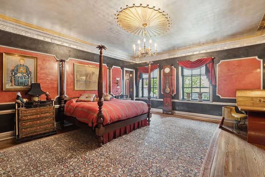 This opulent primary bedroom features a grand four-poster bed as the centerpiece, set against walls with a rich red and black decorative finish. The room is adorned with a large, intricate ceiling medallion, a classic grandfather clock, and a patterned area rug that covers the wood flooring. Large windows with red valances allow natural light to fill the space, which also includes a dedicated desk area and ornate framed artwork.