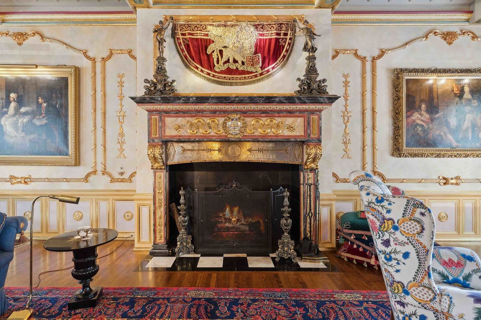 This opulent living room features a grand, ornate fireplace as its central focal point, adorned with intricate gold-leaf detailing and a prominent heraldic crest. The room is framed by elegant wall moldings, two classical oil paintings, and a patterned area rug that anchors the space. A floral-upholstered armchair and a small side table with a lamp complete the sophisticated, traditional aesthetic of this formal sitting area.