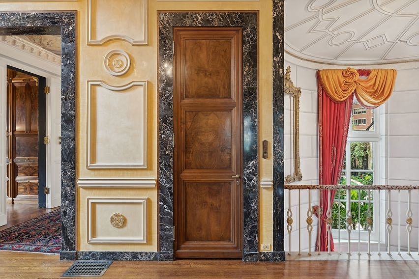 This elegant hallway features a central wooden door framed by a dark, polished stone surround, set against walls adorned with decorative molding and circular medallions. To the right, a curved staircase with a metal railing leads toward a window draped in layered red and gold curtains, while a glimpse into an adjacent room reveals rich wood paneling. The perspective is centered and symmetrical, emphasizing the classical architectural details and the sophisticated, formal aesthetic of the home.