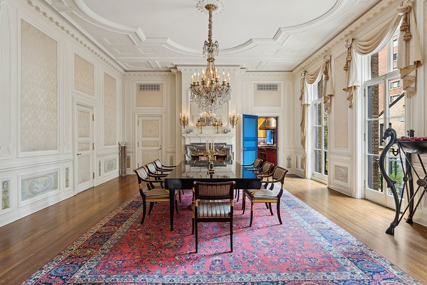 This formal dining room exudes elegance with its ornate crown molding, paneled walls, and a large crystal chandelier centered above a dark, polished dining table. A vibrant, patterned area rug anchors the space, which is furnished with classic striped chairs and features tall windows draped in soft fabric treatments. The room is framed by a striking blue door leading to an adjacent space, creating a sophisticated and grand atmosphere.