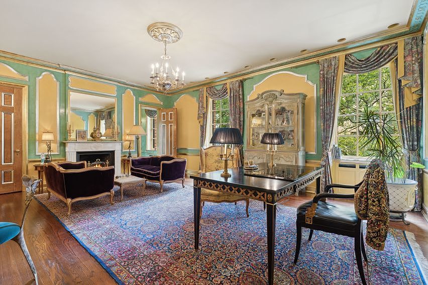 This elegant, traditional-style study features a rich, patterned area rug covering hardwood floors, complemented by walls painted in soft green with gold-trimmed molding. The room is furnished with a dark wood desk, a velvet-upholstered sofa, and a fireplace with a decorative mantel, all illuminated by a classic chandelier and table lamps. Large windows with patterned drapery allow natural light to filter in, creating a sophisticated and scholarly atmosphere.