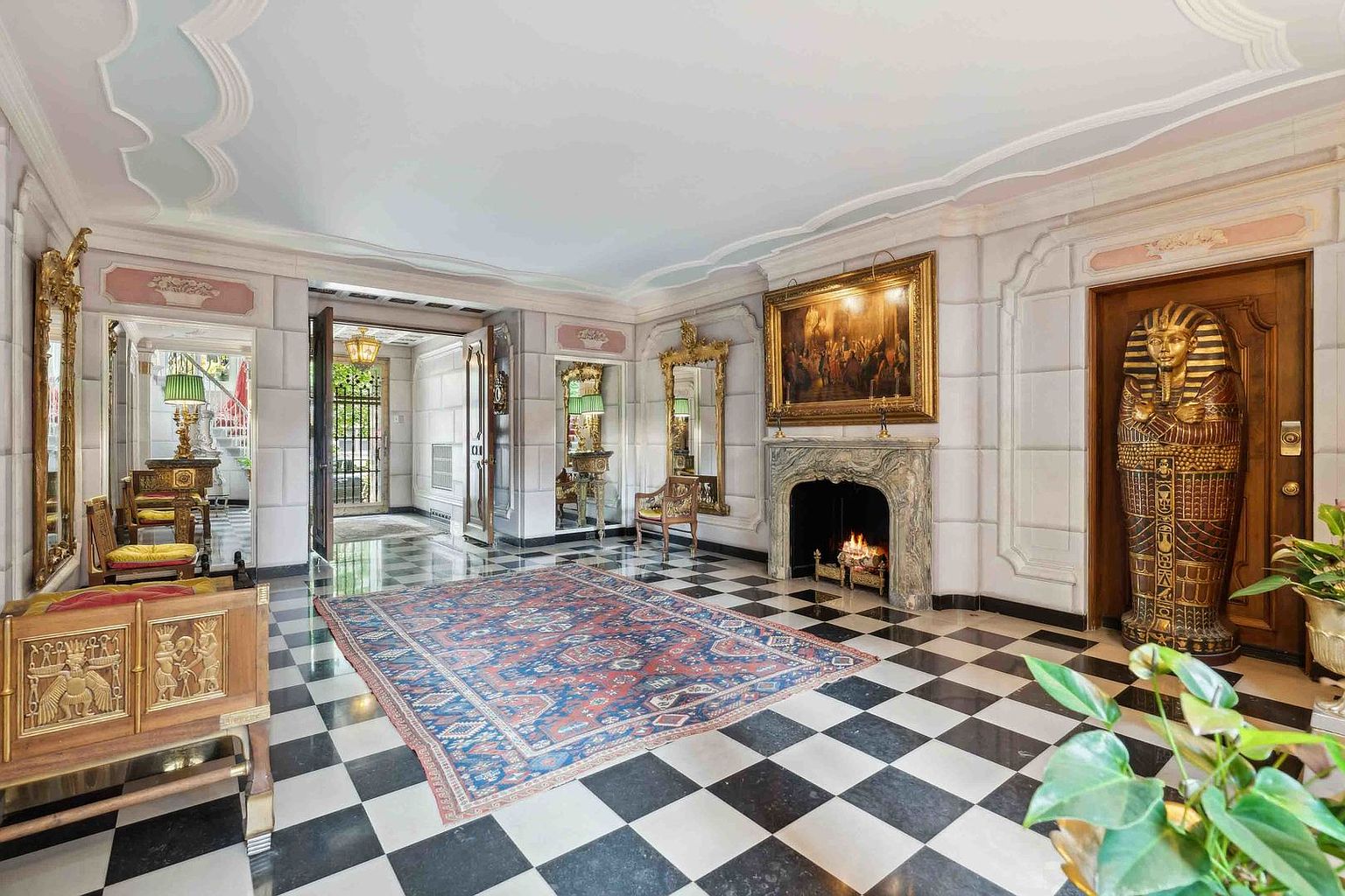 This opulent living room features a striking black-and-white checkered floor and an ornate, eclectic design aesthetic. A large, traditional rug centers the room in front of a stone fireplace with a lit fire, while a prominent Egyptian-style sarcophagus stands near a wooden door. The space is accented with gilded mirrors, classical artwork, and decorative furniture, creating a dramatic and highly stylized atmosphere.