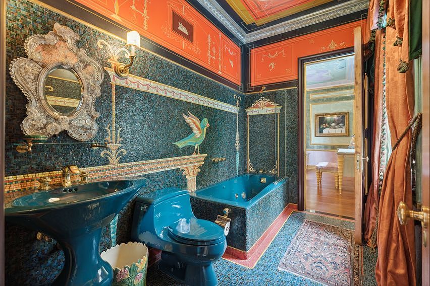 This highly ornate bathroom features deep teal mosaic tile walls adorned with a decorative bird motif and a matching teal pedestal sink and toilet. A dramatic, intricately carved silver-toned mirror hangs above the sink, complemented by a vibrant orange upper wall section with classical-style painted detailing. The space includes a built-in soaking tub with a glass partition and is finished with a patterned area rug, creating a bold, maximalist aesthetic.