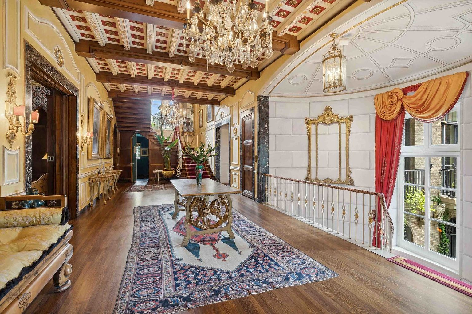 This grand, opulent hallway features rich wood flooring, a coffered ceiling with exposed beams, and an ornate crystal chandelier. A central console table sits atop a patterned area rug, while a staircase leads to an upper level in the background. To the right, a large arched window with elegant drapery allows natural light to illuminate the space, which is accented by a decorative gold-framed mirror on the wall.