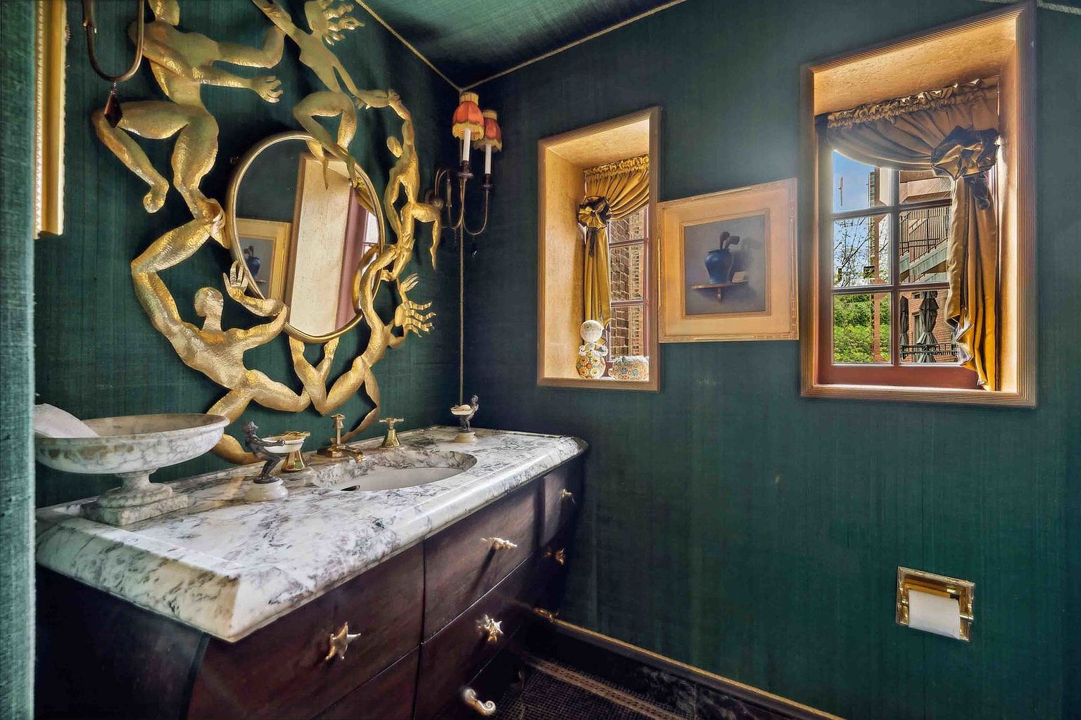 This opulent powder room features deep green textured walls and a striking gold-toned sculptural mirror depicting figures surrounding the glass. A dark wood vanity topped with a patterned stone surface and a vessel sink serves as the focal point, complemented by ornate gold hardware and wall-mounted light fixtures. Two recessed windows with gold-toned curtains provide natural light, while a framed piece of art hangs between them, contributing to the room's dramatic and sophisticated aesthetic.