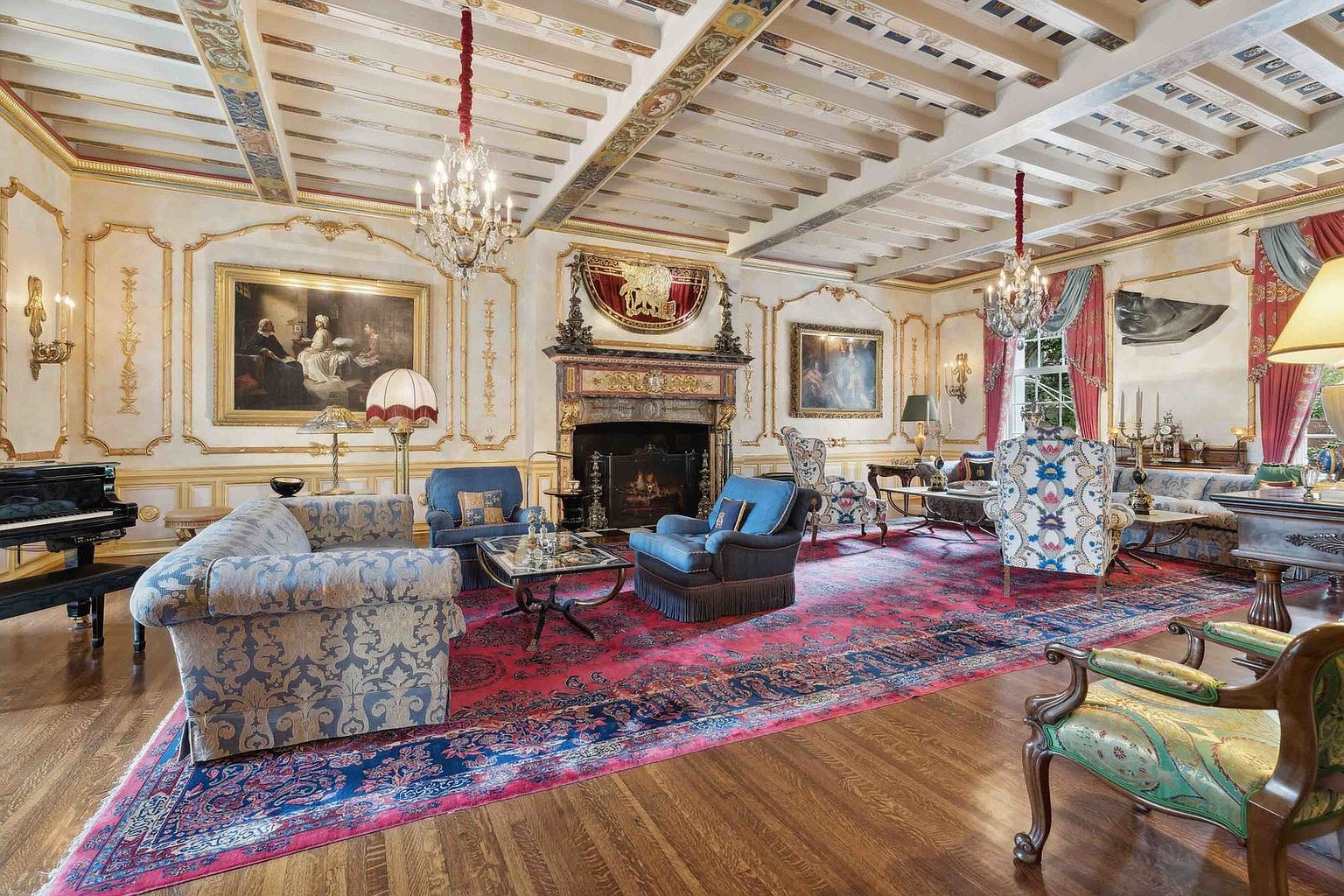 This opulent living room features a grand, formal aesthetic with intricate crown molding, a coffered ceiling with painted details, and a large, ornate fireplace as a central focal point. The space is furnished with a mix of classic upholstered sofas and armchairs arranged on a large, vibrant red patterned area rug over hardwood floors. Two crystal chandeliers hang from the ceiling, and the room is decorated with fine art, gold-trimmed wall panels, and heavy drapery, creating a sophisticated and historic atmosphere.