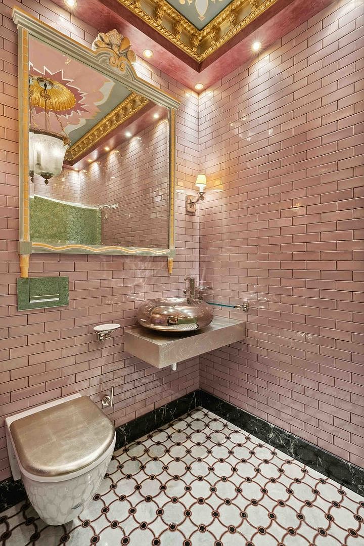 This ornate powder room features dusty rose subway-style wall tiles and a decorative ceiling with gold-leaf detailing. A metallic vessel sink sits atop a floating stone vanity, complemented by a large, elaborately framed mirror and a patterned tile floor with a dark stone border. The space is illuminated by a combination of recessed lighting and a wall-mounted sconce, creating a luxurious and dramatic atmosphere.