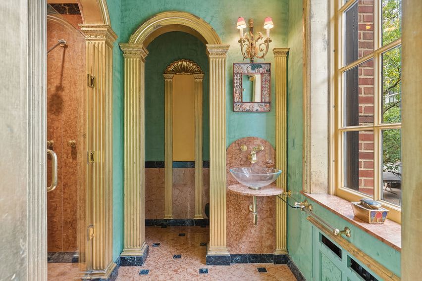 This ornate bathroom features a distinct neoclassical design with vibrant green walls, gold-painted columns, and a pink stone floor. A glass vessel sink is mounted on a pink stone pedestal, complemented by a decorative wall sconce and a small framed mirror. The space includes a glass-enclosed shower area and a large window that allows natural light to illuminate the intricate architectural details.