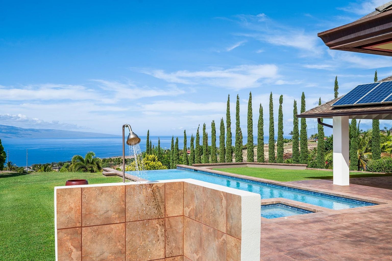 This image showcases a luxurious outdoor pool and spa area with stunning ocean views. The pool features a unique design with a built-in spa and an outdoor shower, all surrounded by well-maintained landscaping and tall, slender trees. The presence of solar panels on the nearby structure adds an eco-friendly touch to this upscale property.